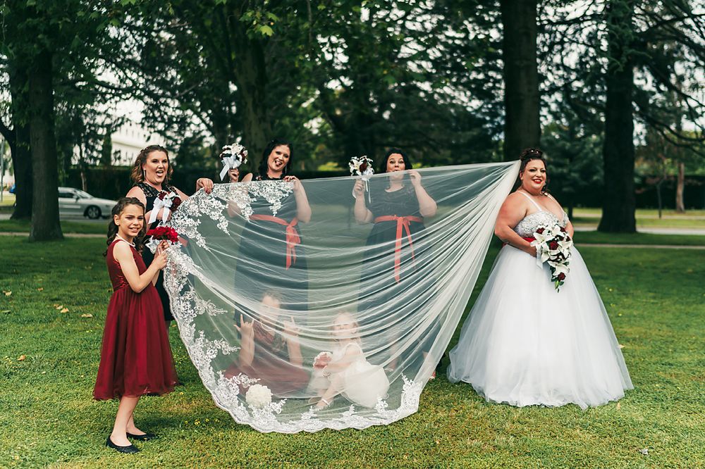 A group in white and burgundy dresses plays with a flowing veil in a lush green outdoor park setting.