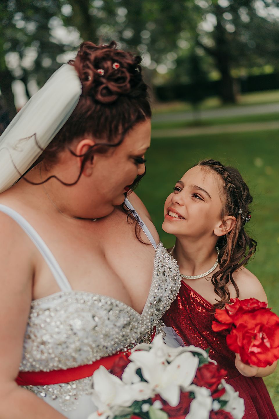 A bride in a sparkly white dress with red accents holds a bouquet while sharing a tender moment in an outdoor setting.