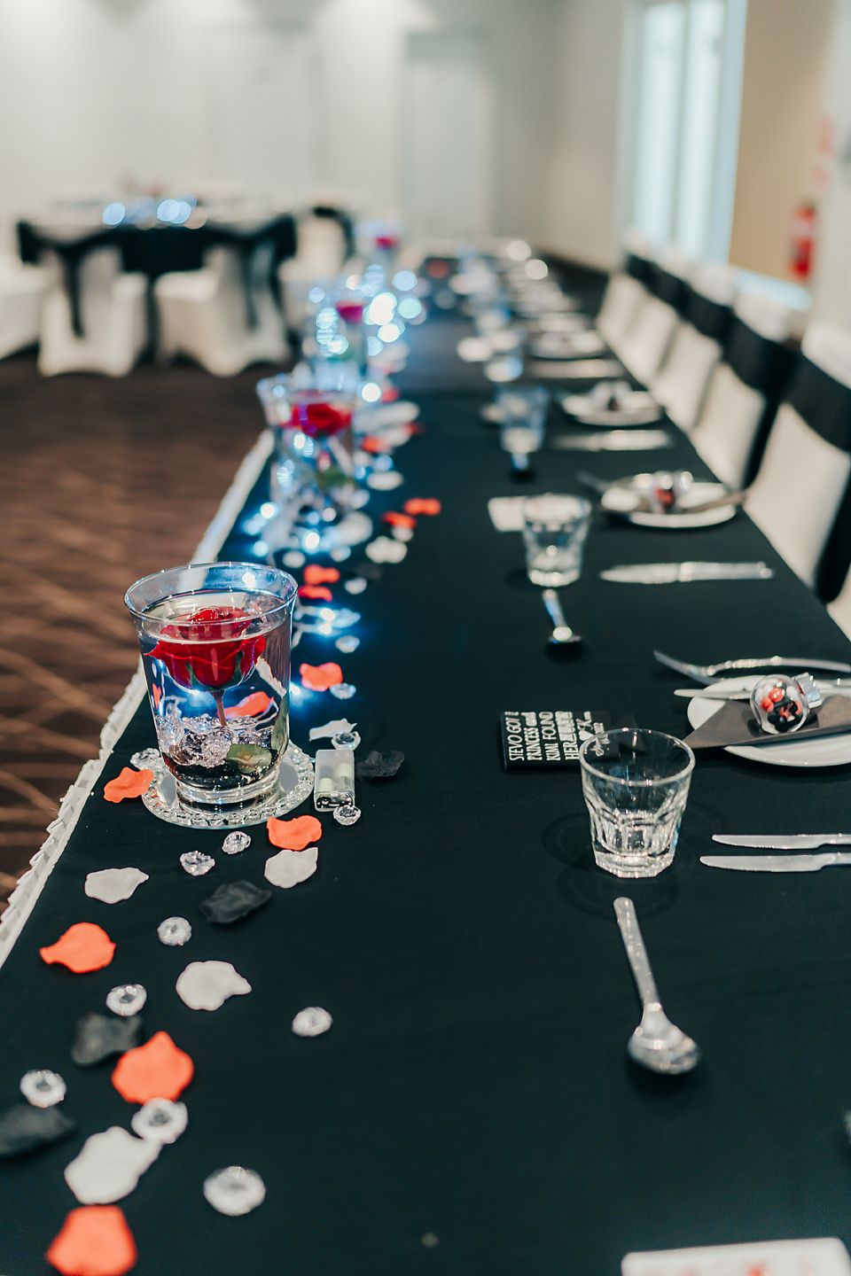 An elegant wedding reception table with black linens decorated with red drinks, scattered petals and formal place settings.