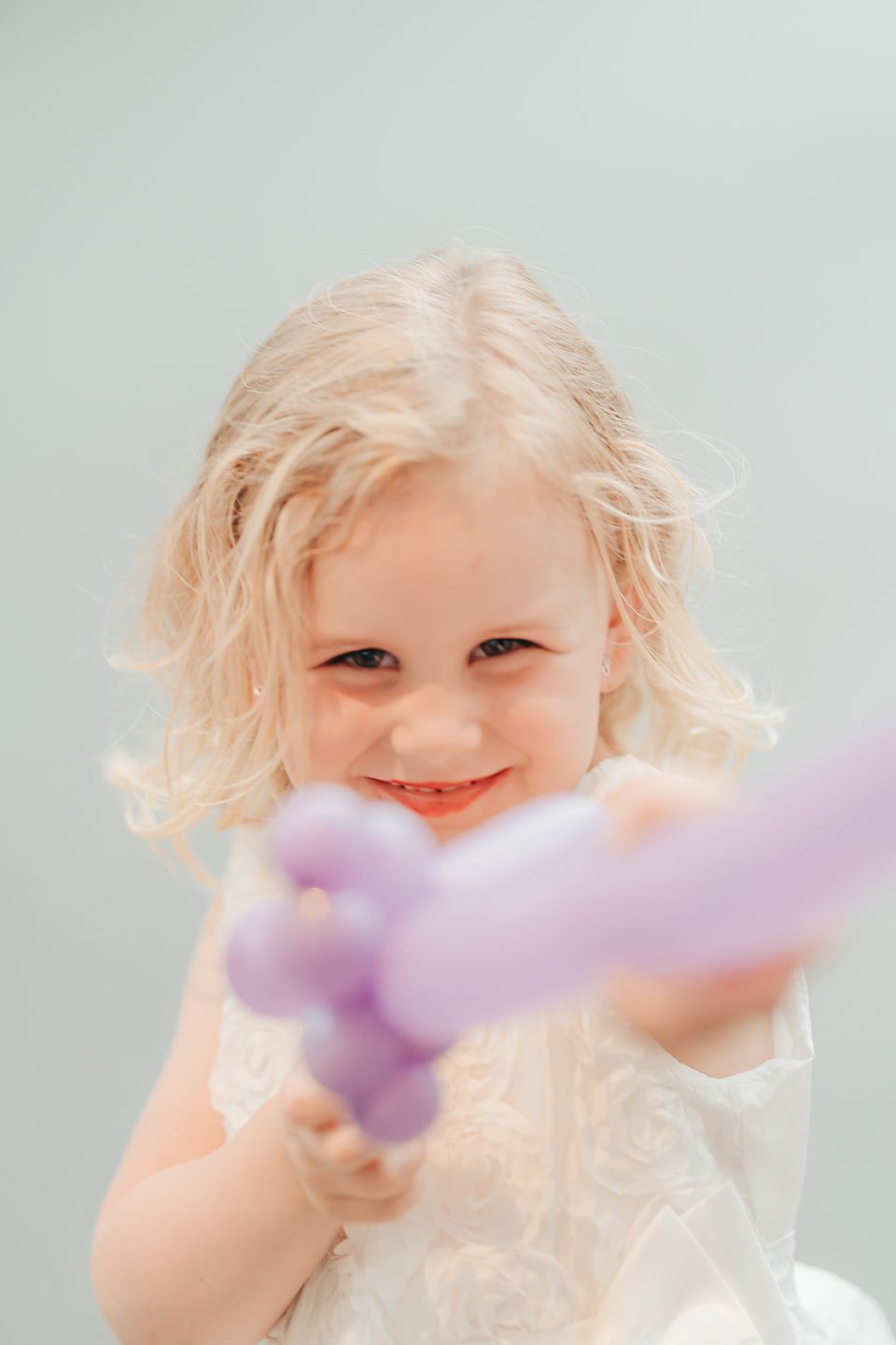 Child with light blonde hair holding a purple toy and wearing white, smiling joyfully against a light background.