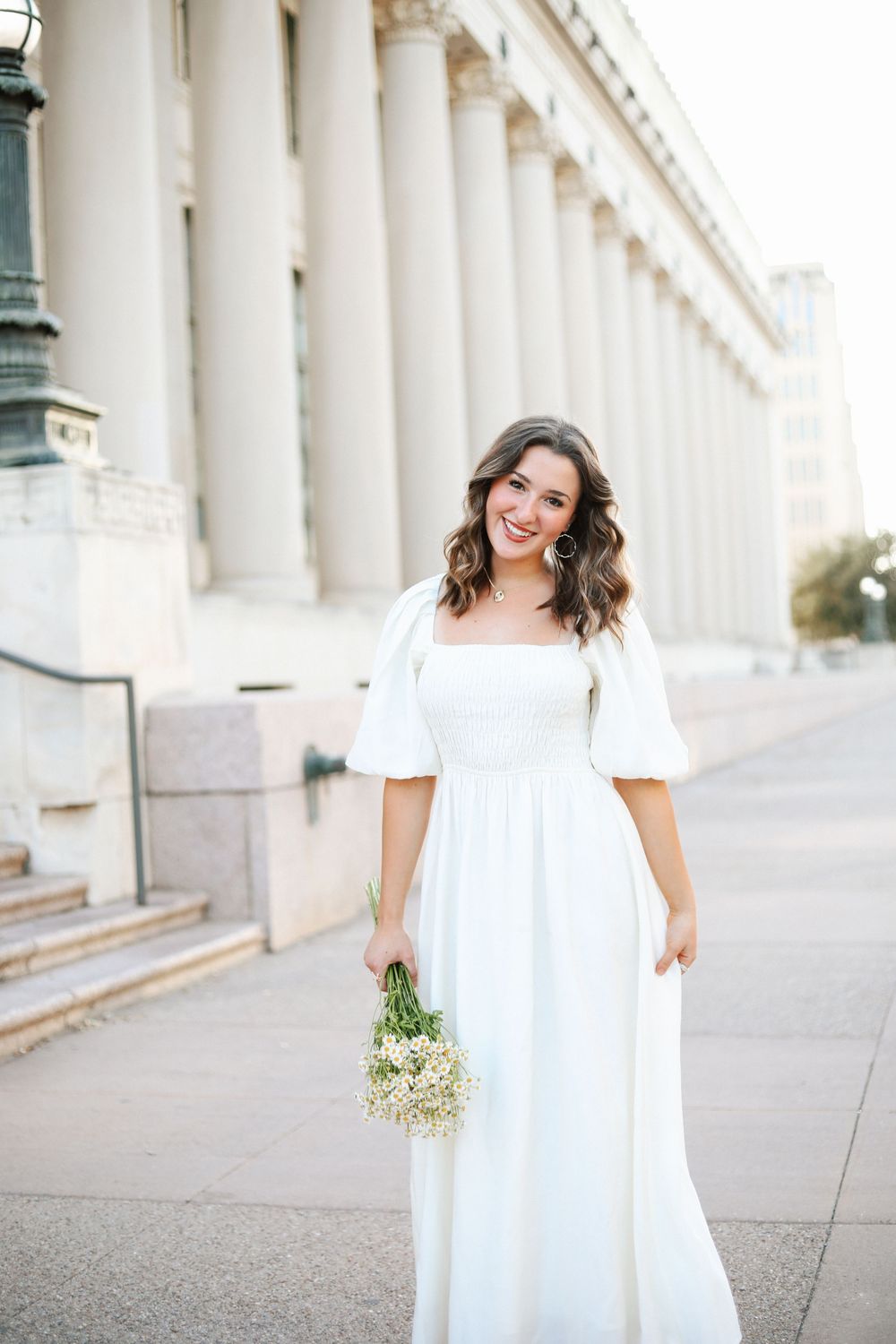 A woman in a white dress poses elegantly near classical columns while holding a bouquet of baby's breath flowers.