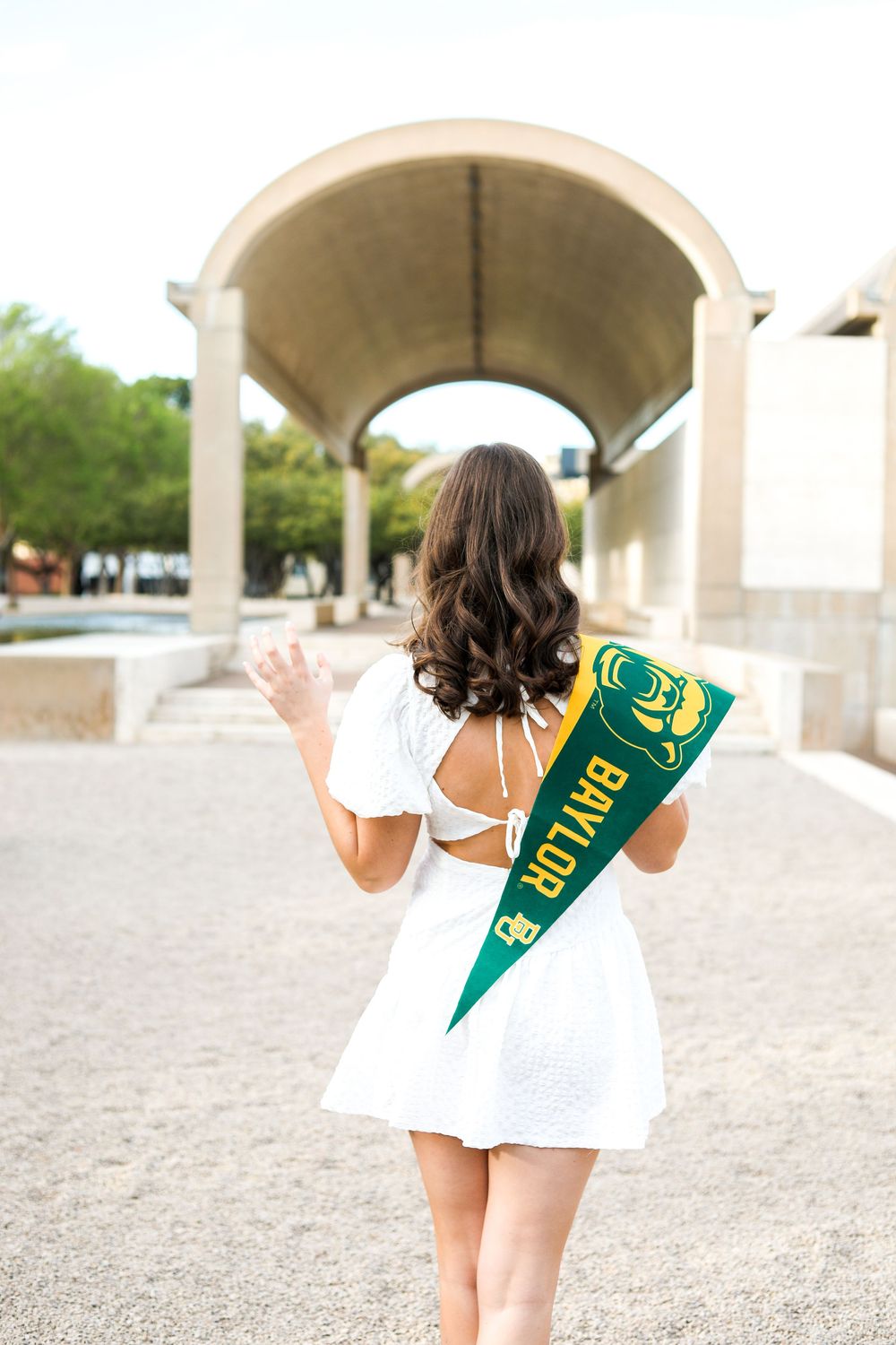 A person in white dress holds a Baylor University pennant while walking under an archway.