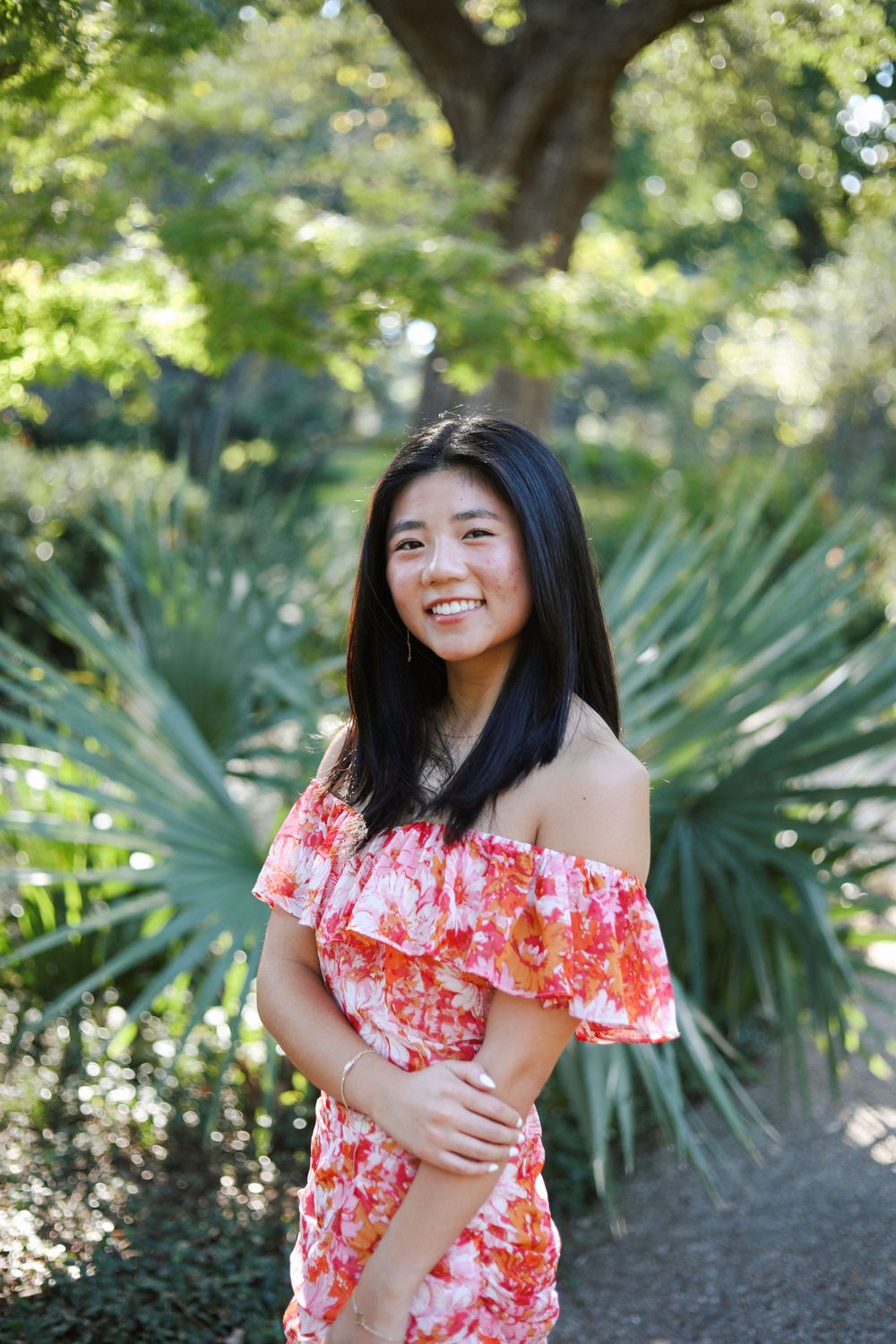 A woman wearing an off-shoulder floral dress stands in a lush garden setting with palm fronds.