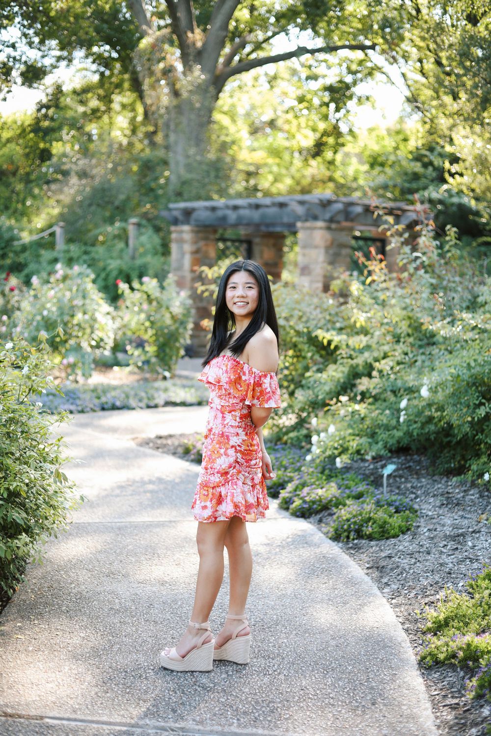 A woman in a red floral dress poses on a curved garden path surrounded by greenery.