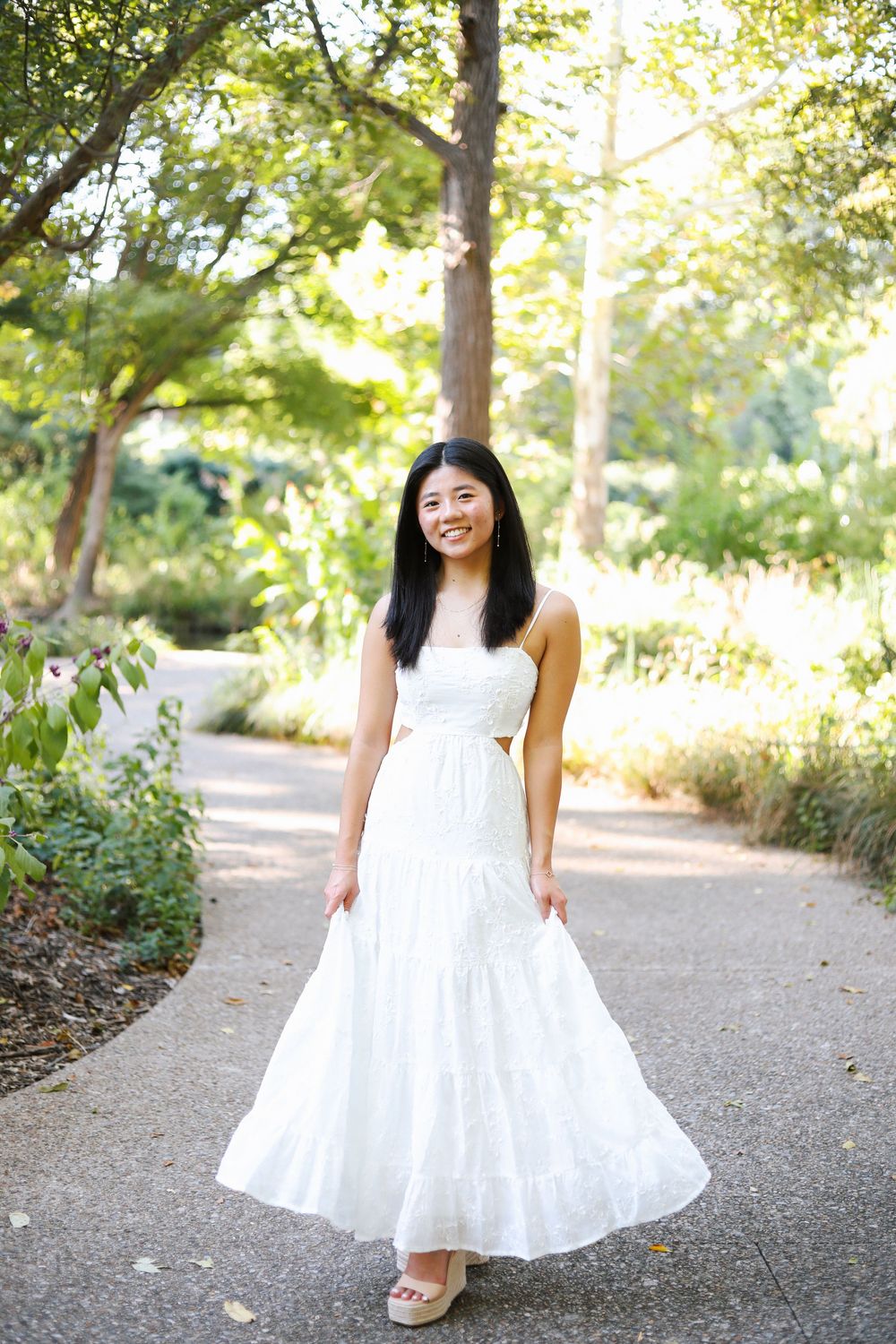 A woman in a flowing white maxi dress stands on a garden path surrounded by trees and sunlight.