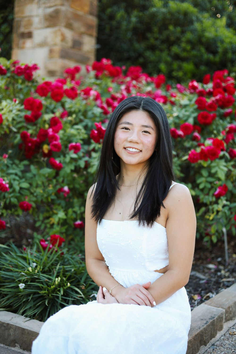 A woman in white dress sits near vibrant red roses in a garden setting.