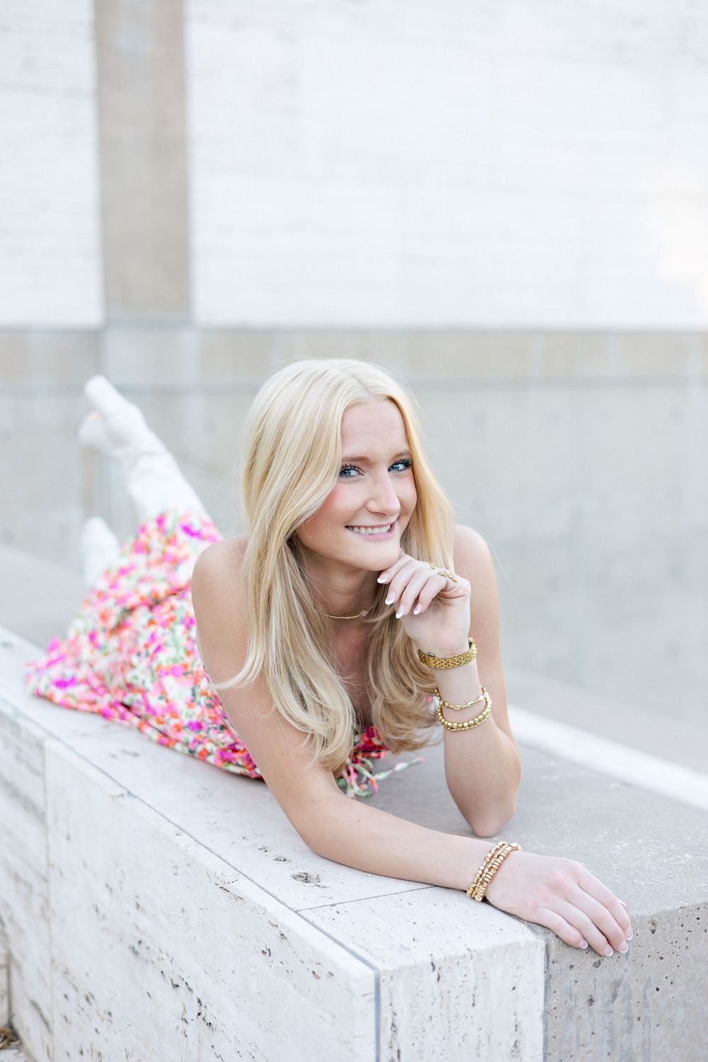 A woman with blonde hair wearing a pink floral dress lies casually on a light colored stone surface outdoors.