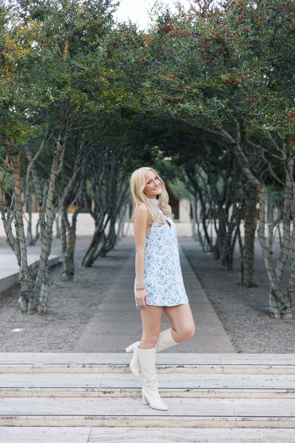 A blue and white floral mini dress paired with white boots stands out against a backdrop of trees forming a natural tunnel.