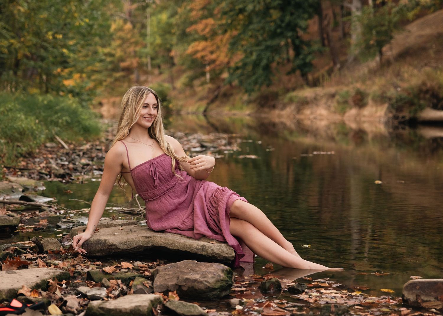 Woman in burgundy dress posing gracefully by a creek with autumn leaves.