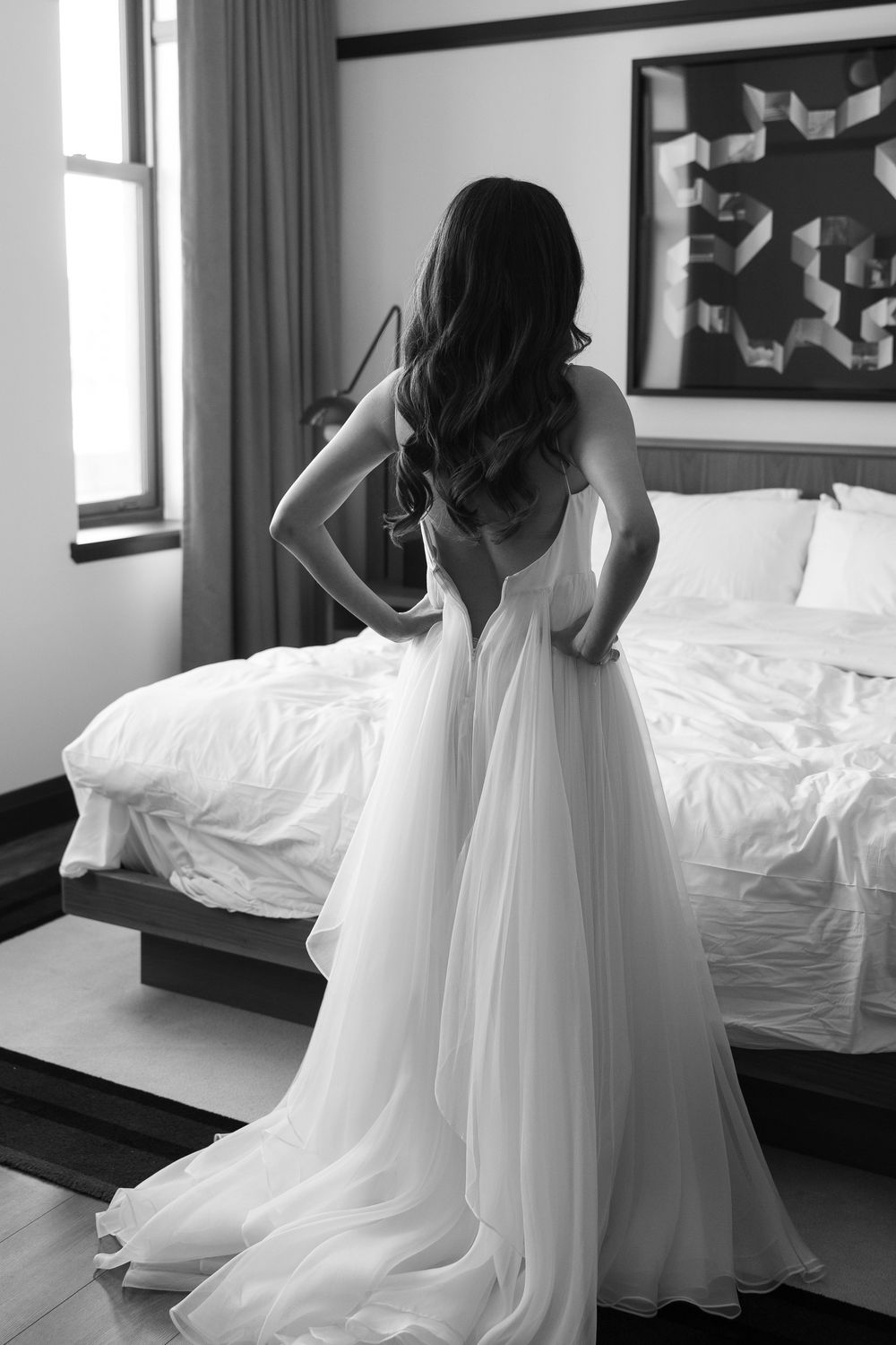 A bride in a flowing white wedding gown stands beside a bed in black and white.