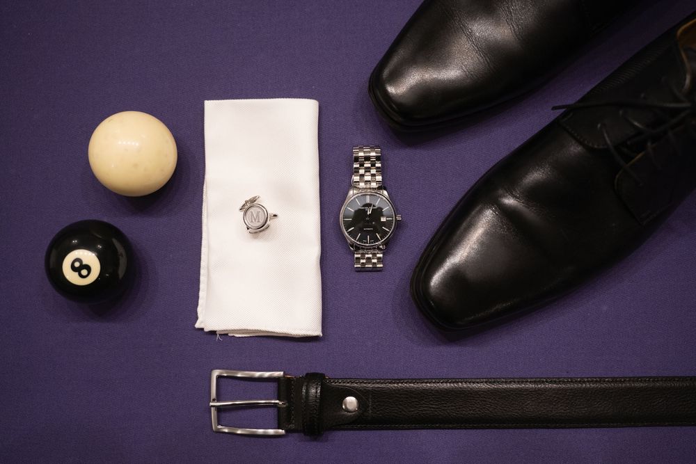 Wedding accessories including black dress shoes, rings, and watch on purple background.