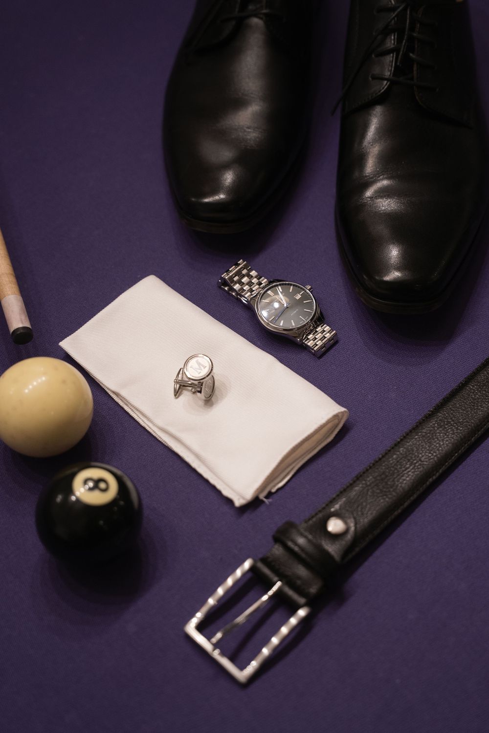 Wedding accessories including black dress shoes, watch, billiard balls, belt and ring on purple background.