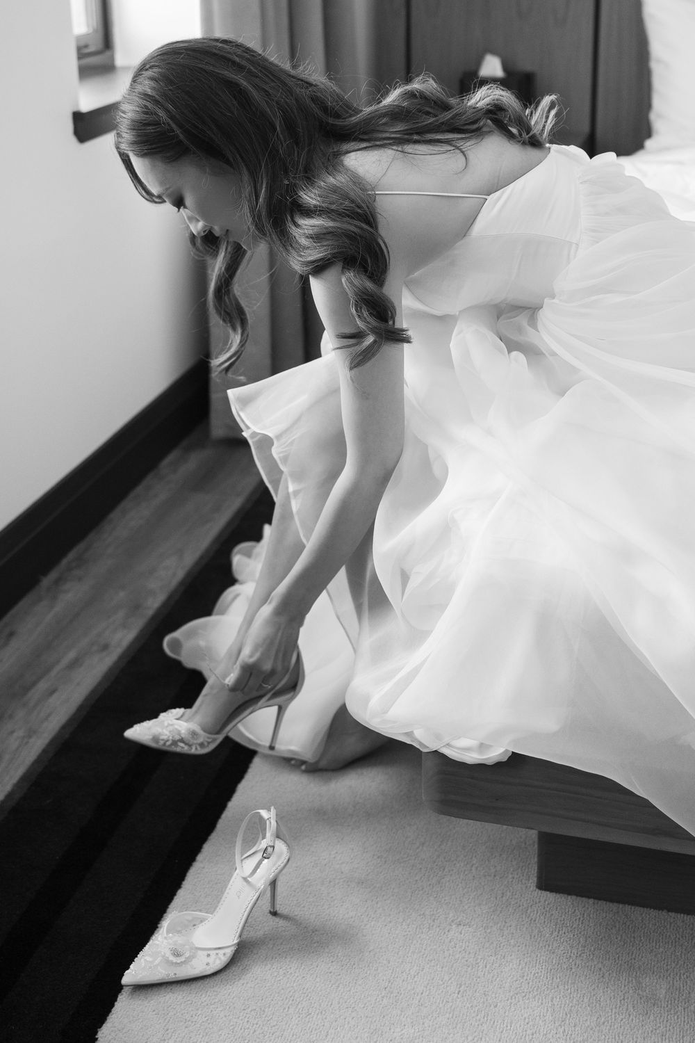 Black and white photo of bride in flowing dress putting on elegant heeled shoes.