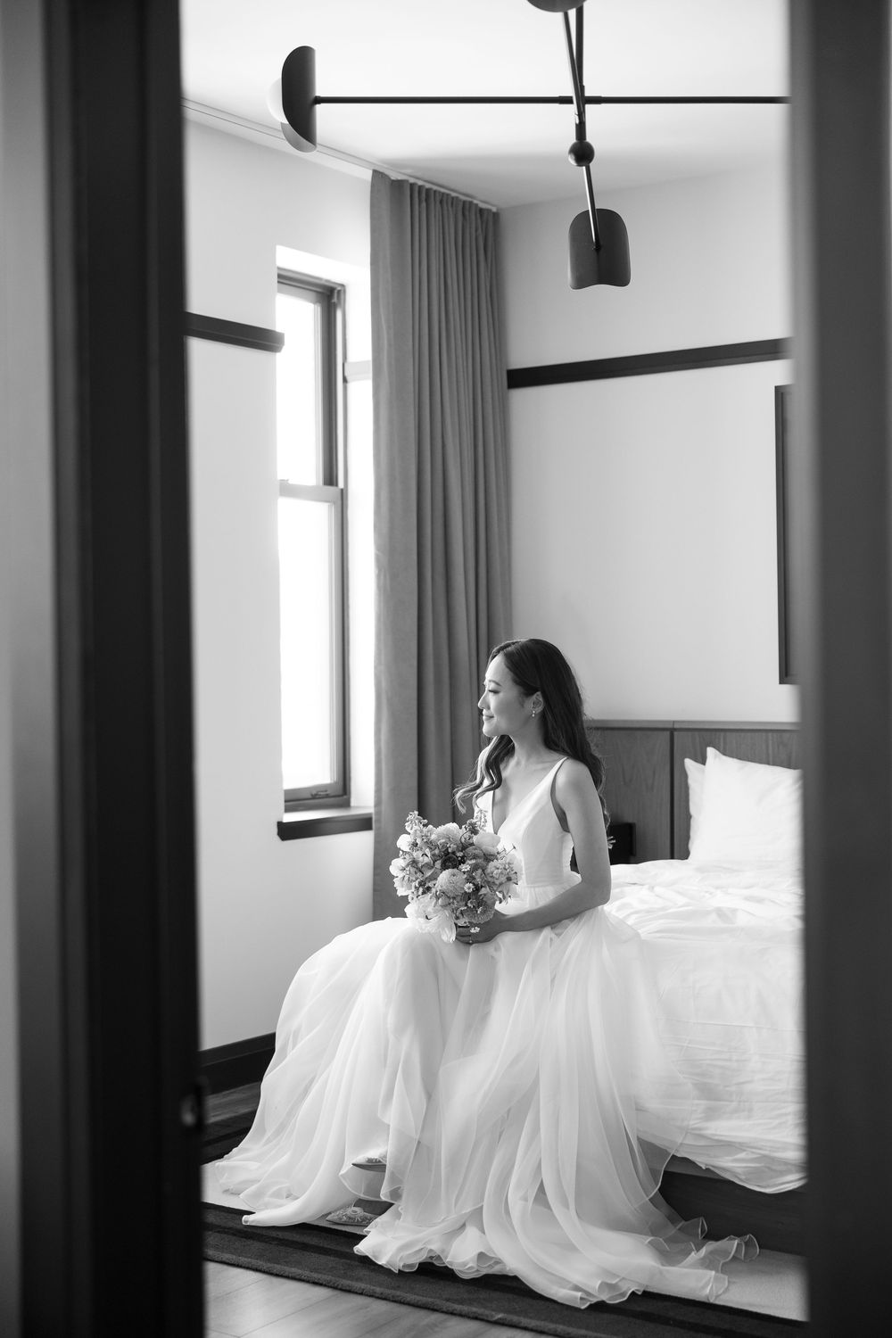 Black and white photograph of a bride wearing a flowing white wedding dress sitting in a modern room with natural light.