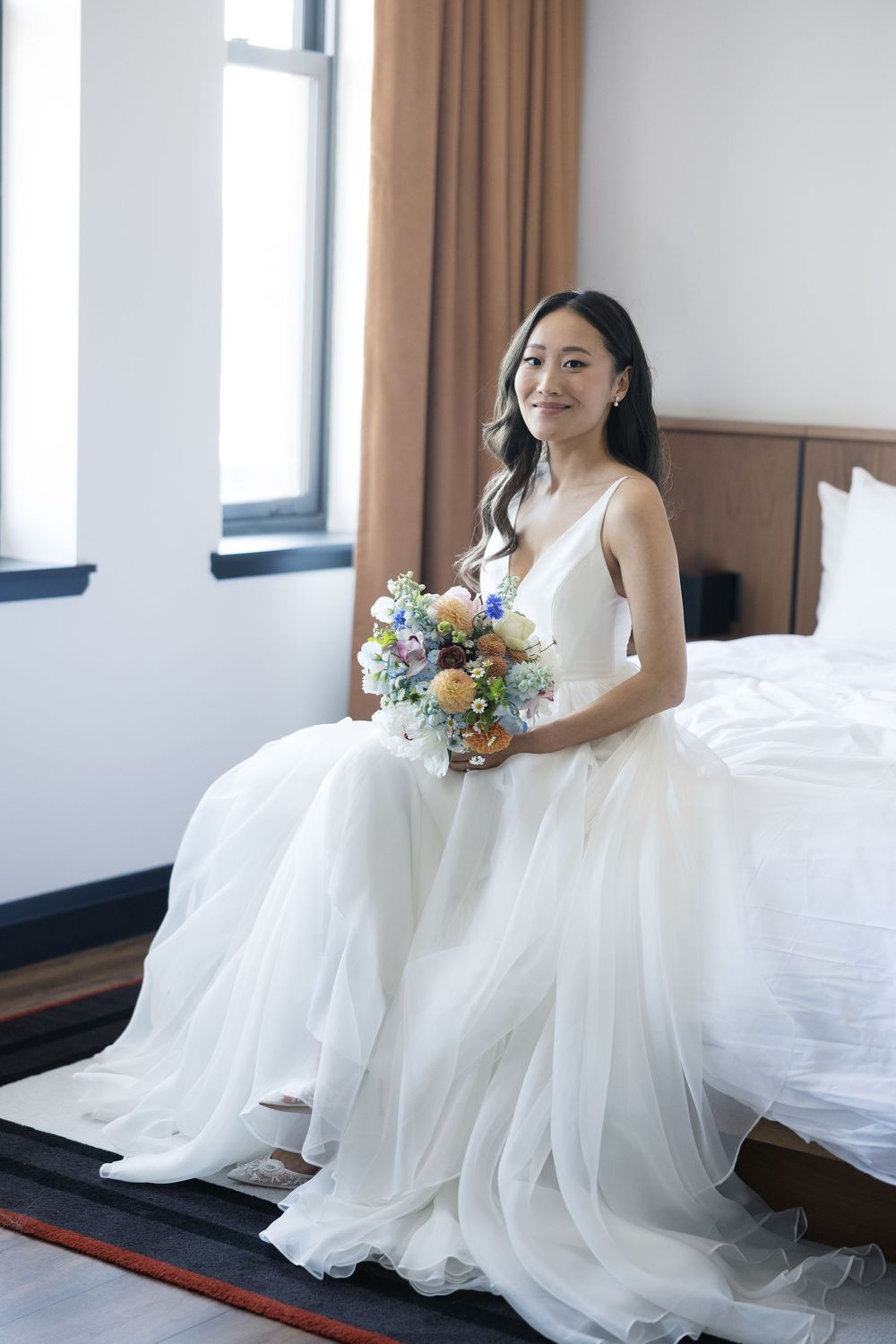 A bride in an elegant white gown sits on the edge of a bed holding a pastel-colored bouquet.