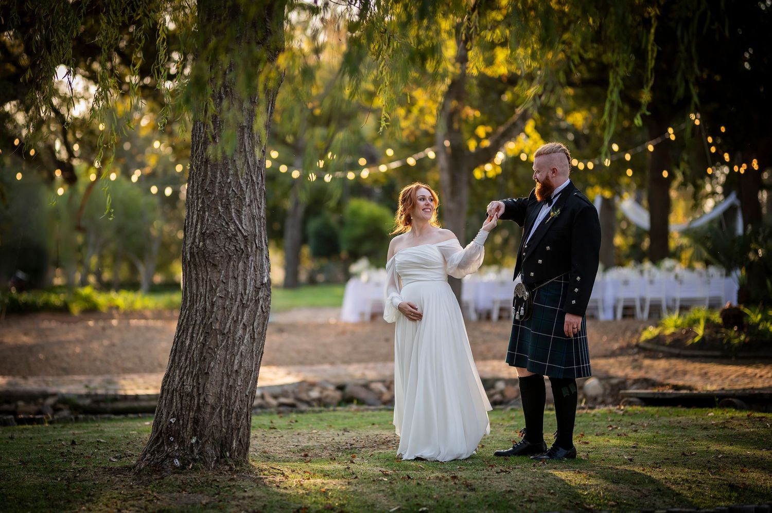 A wedding couple stands beneath a tree at dusk with twinkle lights strung overhead in a garden setting.