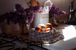 Fresh eggs in a checkered cloth basket with purple lilac flowers in vases on a kitchen counter.