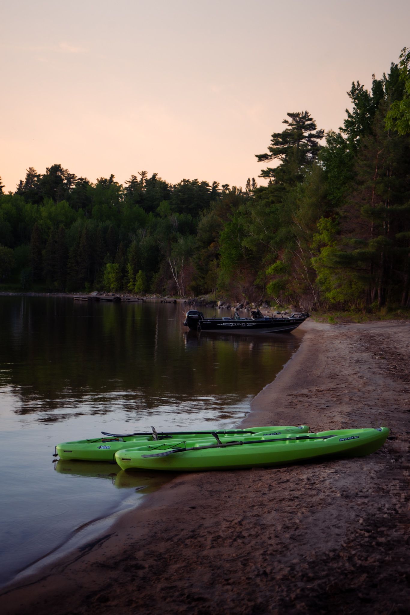 Green kayaks rest on a beach shore at sunset with a larger boat visible in the background.