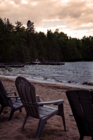 Adirondack chairs on a sandy beach with pine trees and cloudy skies in the background.