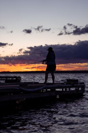 Silhouette of figure fishing on dock during dramatic orange and purple sunset.