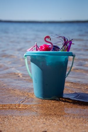 Turquoise beach bucket with pink flamingo toys sitting in wet sand by water.