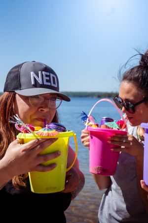 Two friends enjoying colorful summer drinks from festive buckets by the lake on a sunny day.