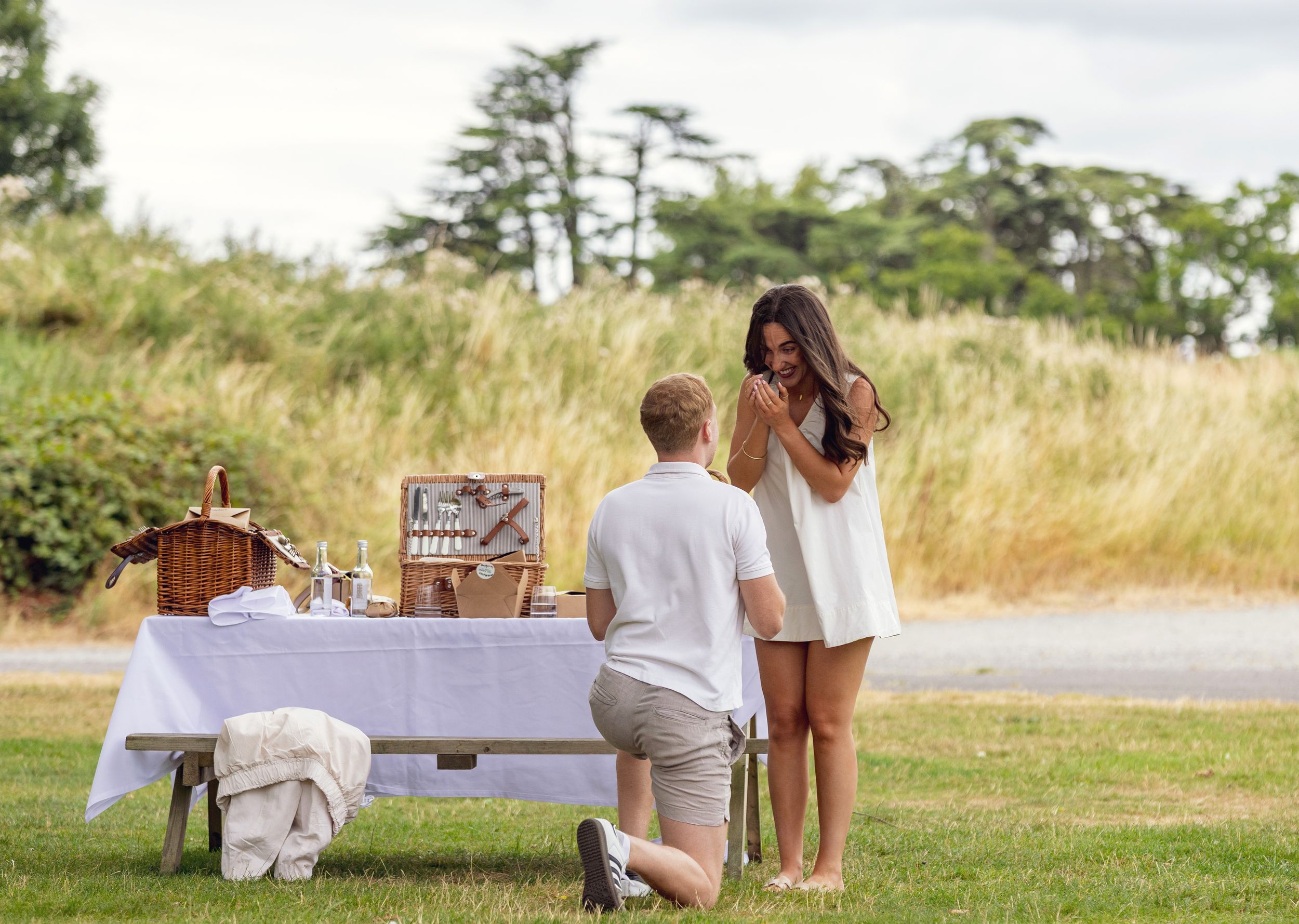 Man proposing on one knee to woman at outdoor picnic setup with white tablecloth and trees in background.
