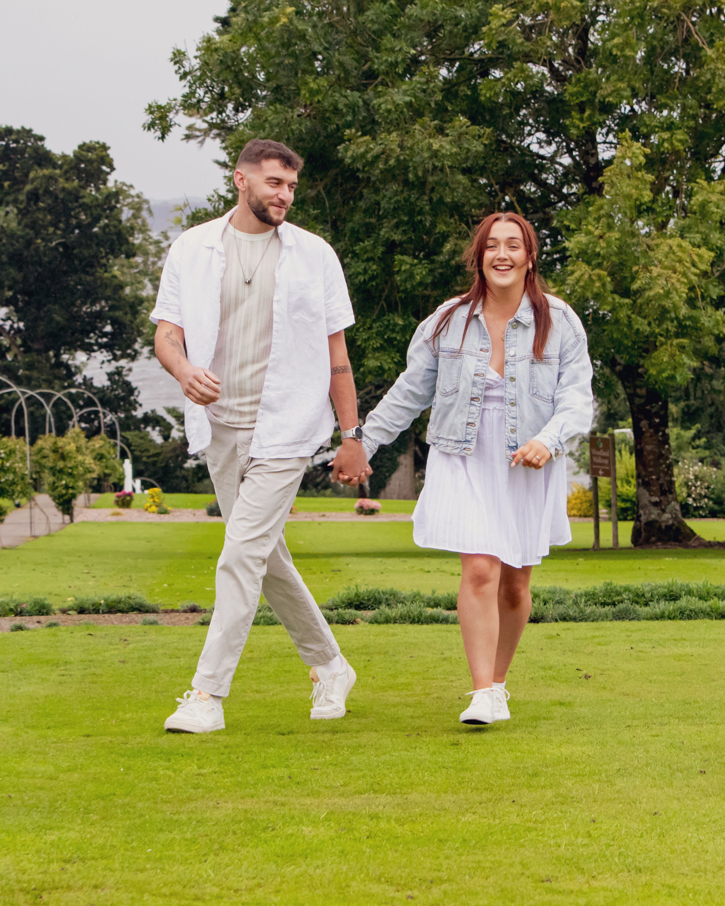 Playful couple in white outfits dancing and twirling together on a lush green lawn with trees in the background.