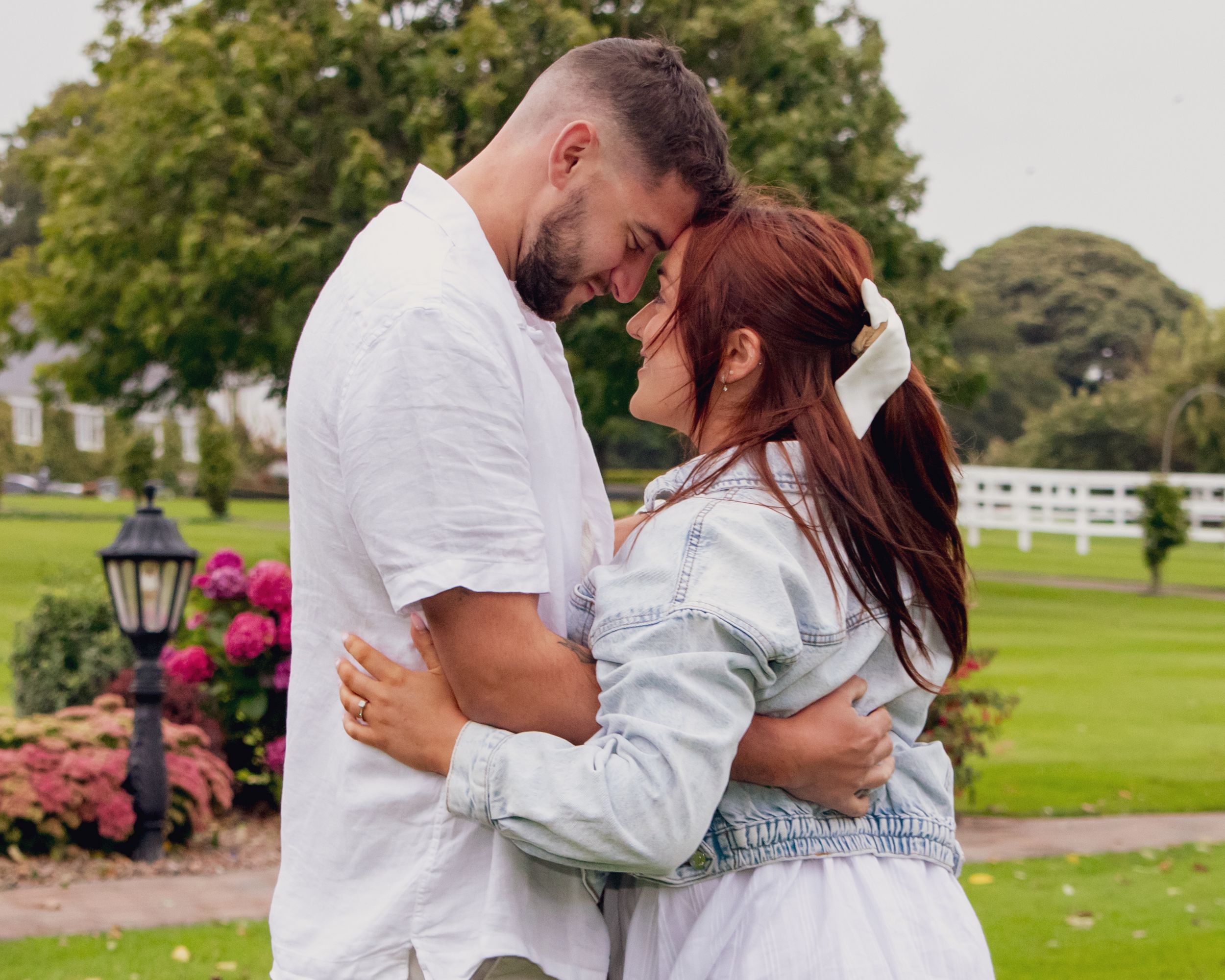 Romantic couple in white and denim outfits sharing intimate moments on a grassy lawn with white fences.