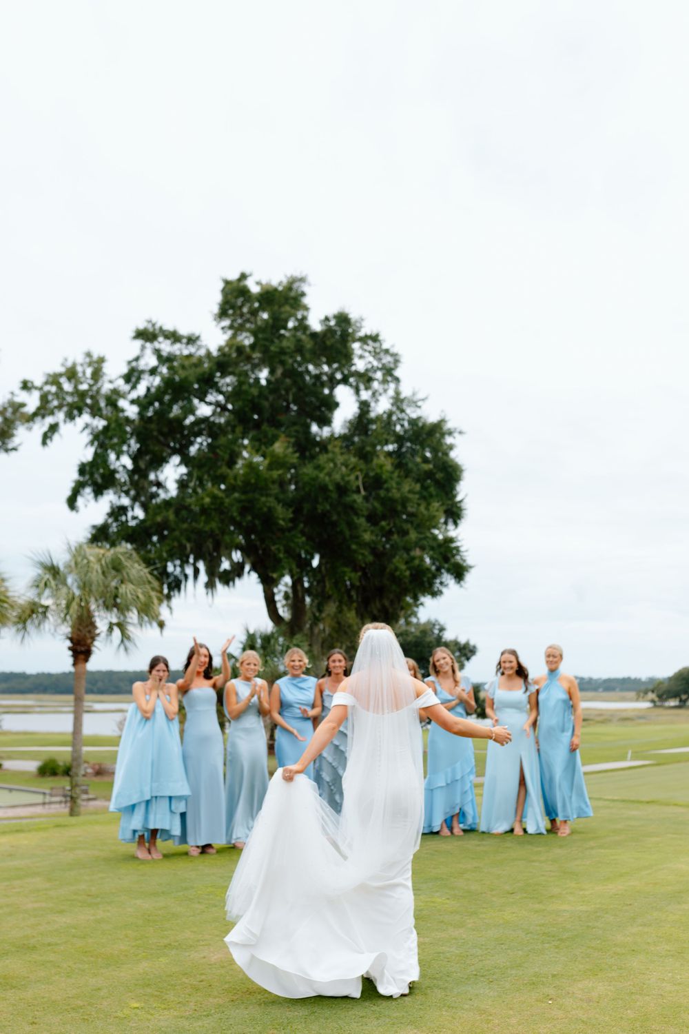 Bride in white gown and veil facing bridesmaids in light blue dresses on green lawn.