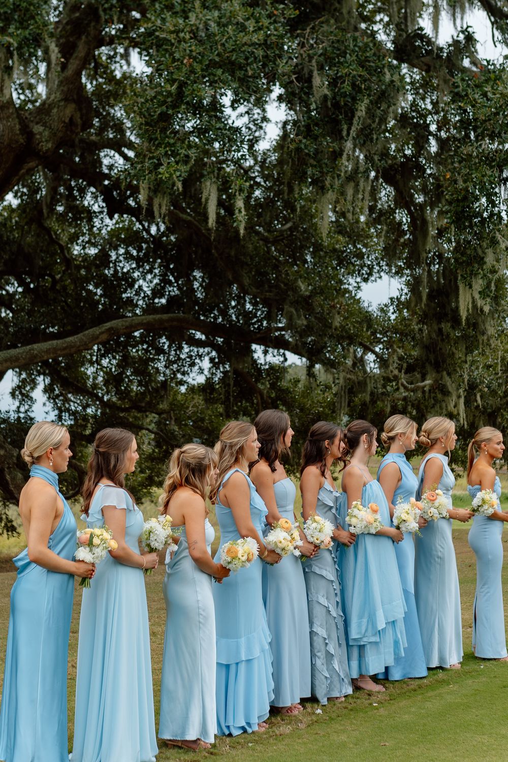 Bridesmaids in matching light blue gowns holding white bouquets stand in a row outdoors beneath a large oak tree.