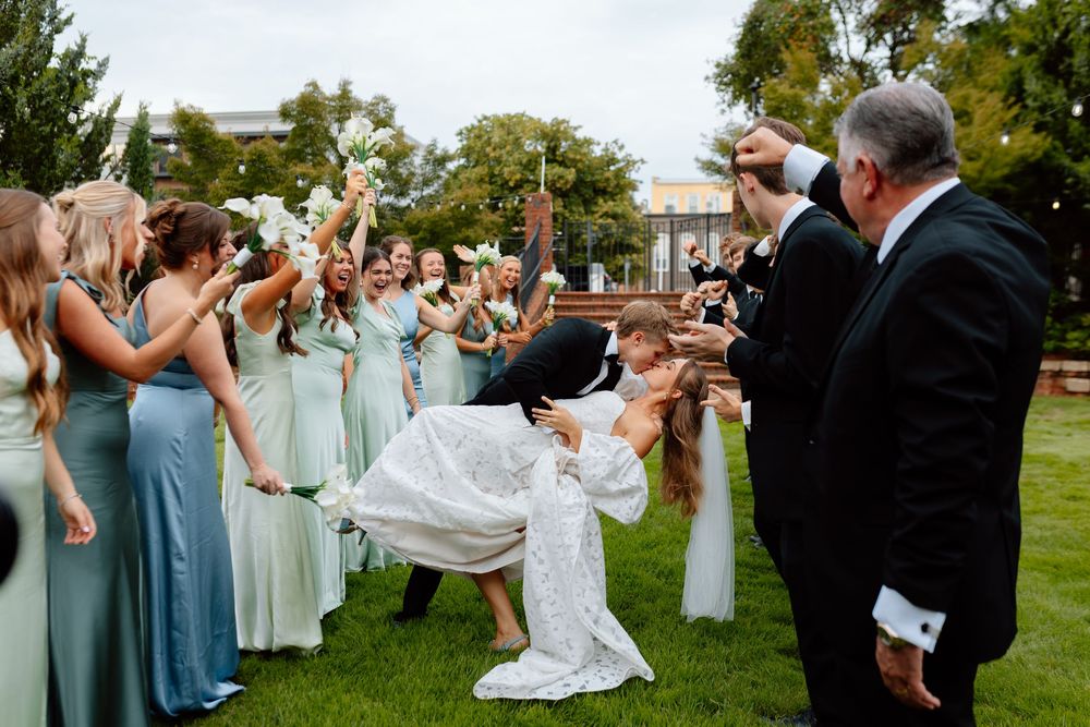 Groom dips bride during wedding exit while bridesmaids in sage green dresses cheer on a green lawn.