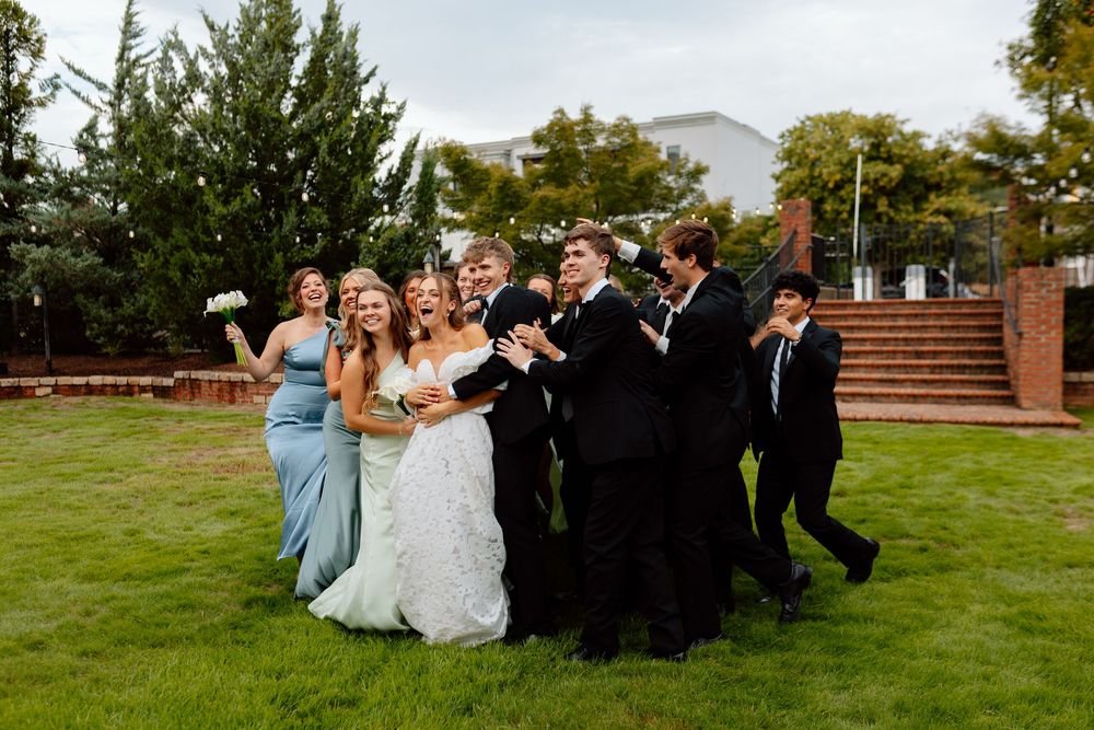 Wedding party celebrating outdoors on green lawn, bride in white gown with bridesmaids in blue and groomsmen in black tuxedos.
