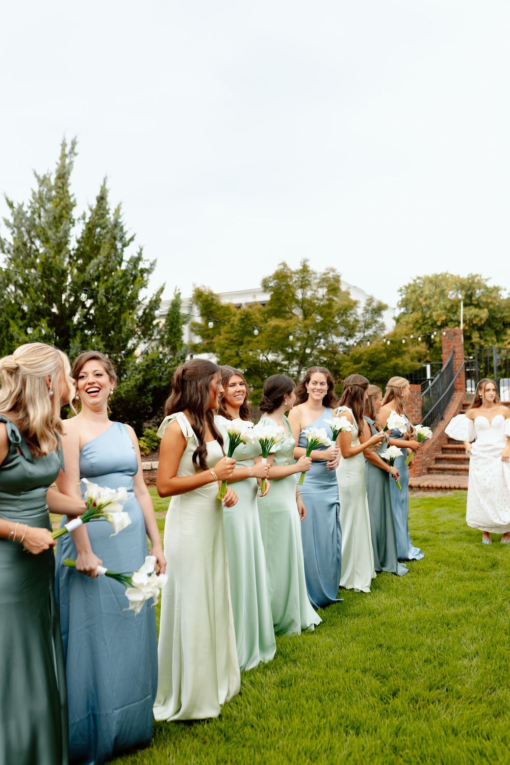 Bridesmaids in blue and mint gowns holding white bouquets line up outdoors on green grass at a wedding.
