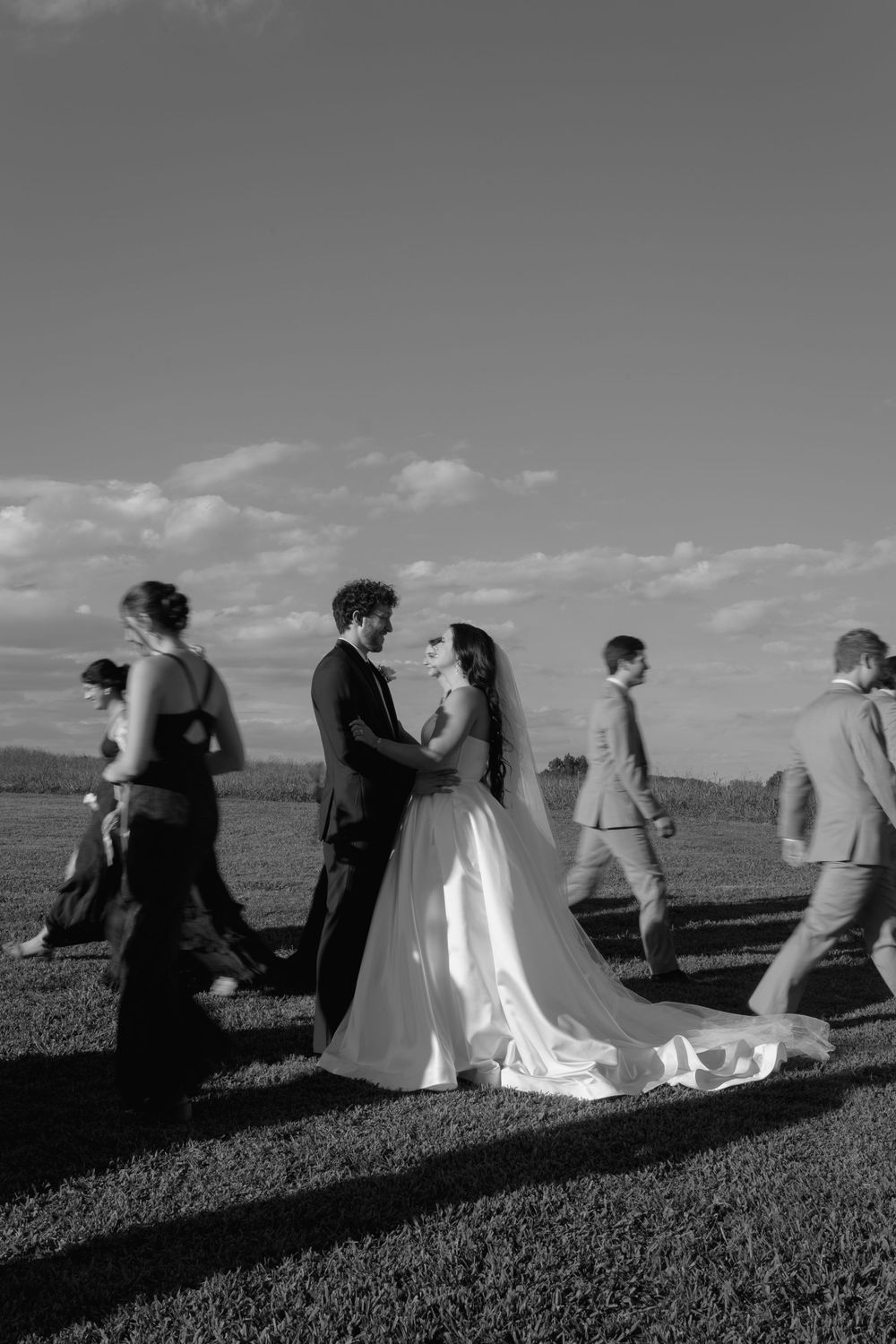 Black and white photo of bride and groom embracing outdoors while wedding party walks past under cloudy sky.
