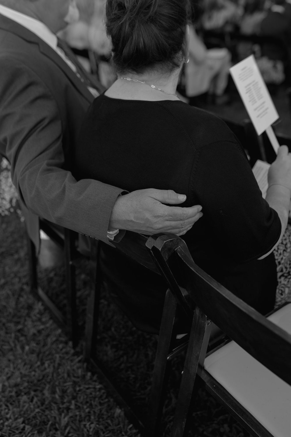 Black and white photo of a man placing his hand on a woman's back while seated at an outdoor ceremony.