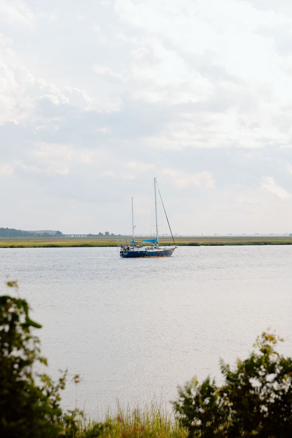 A sailboat anchored in calm coastal waters surrounded by green marshlands under a partly cloudy sky.
