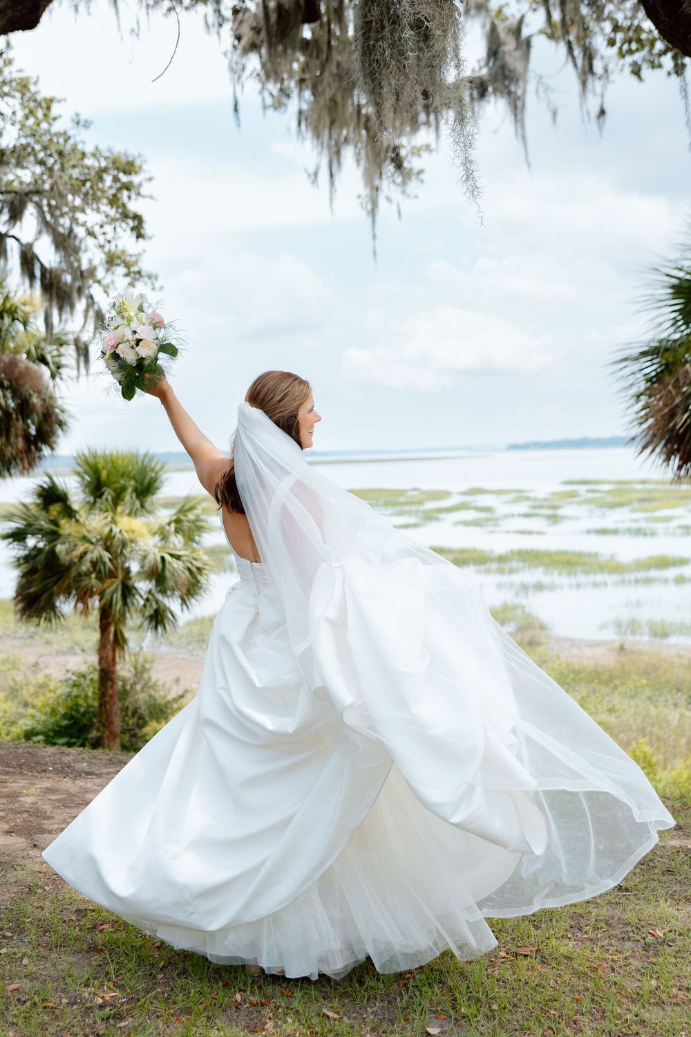 Bride in white gown twirling with bouquet raised, veil flowing, near coastal marsh with Spanish moss trees.