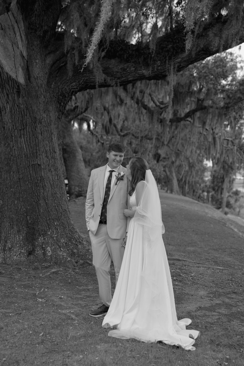 Black and white photo of bride and groom sharing a romantic moment under a large moss-draped oak tree.