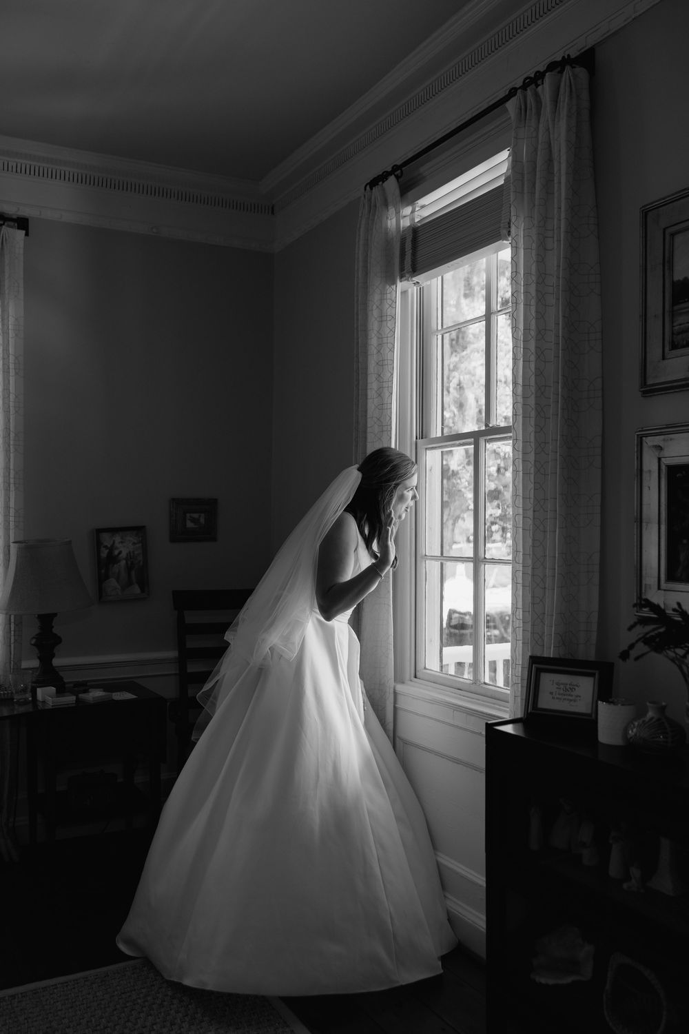 Bride in white gown and veil stands by sunlit window in elegant room, captured in black and white photography.