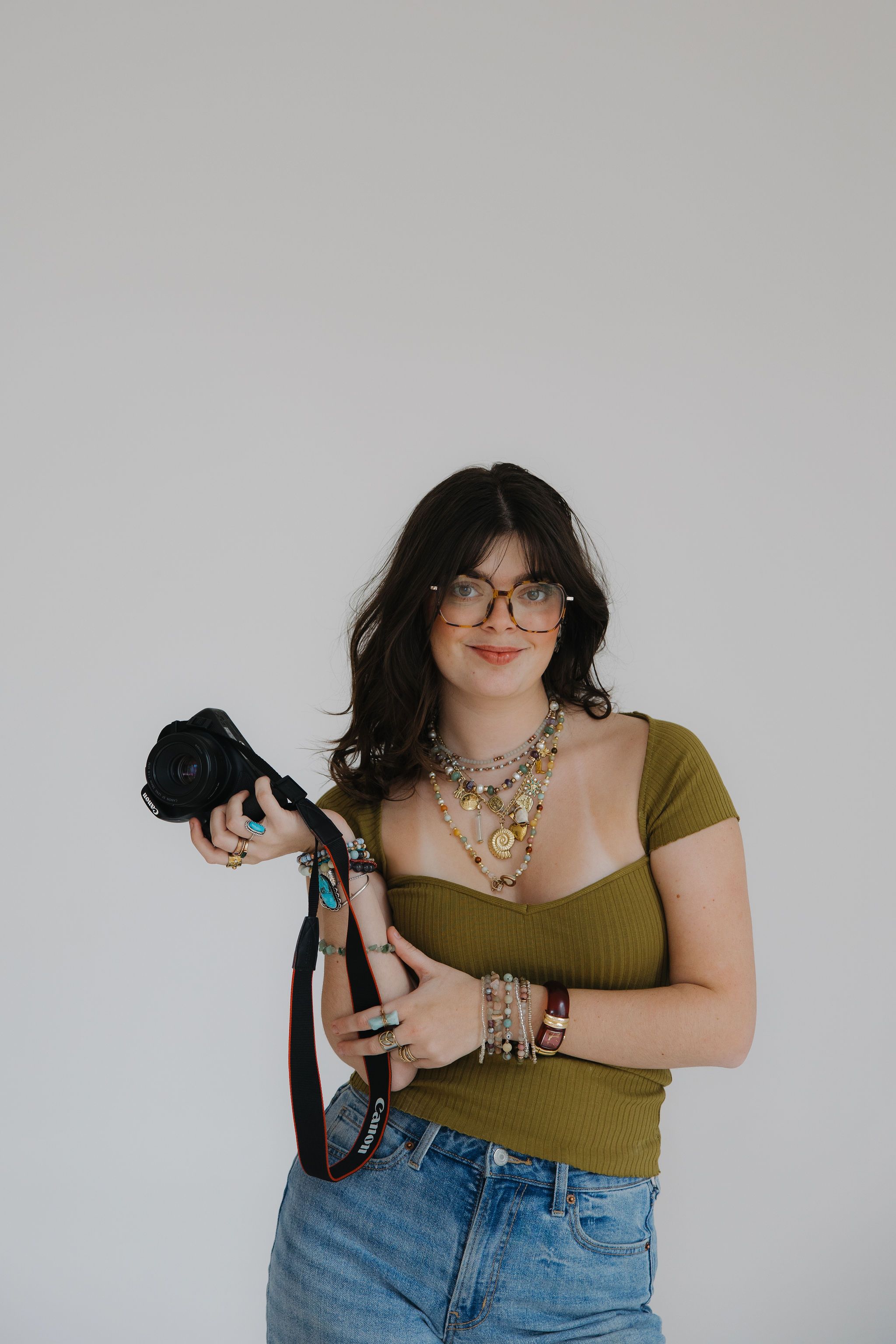 Photographer in olive green top and denim holds professional camera while wearing stylish glasses and jewelry against white wall.