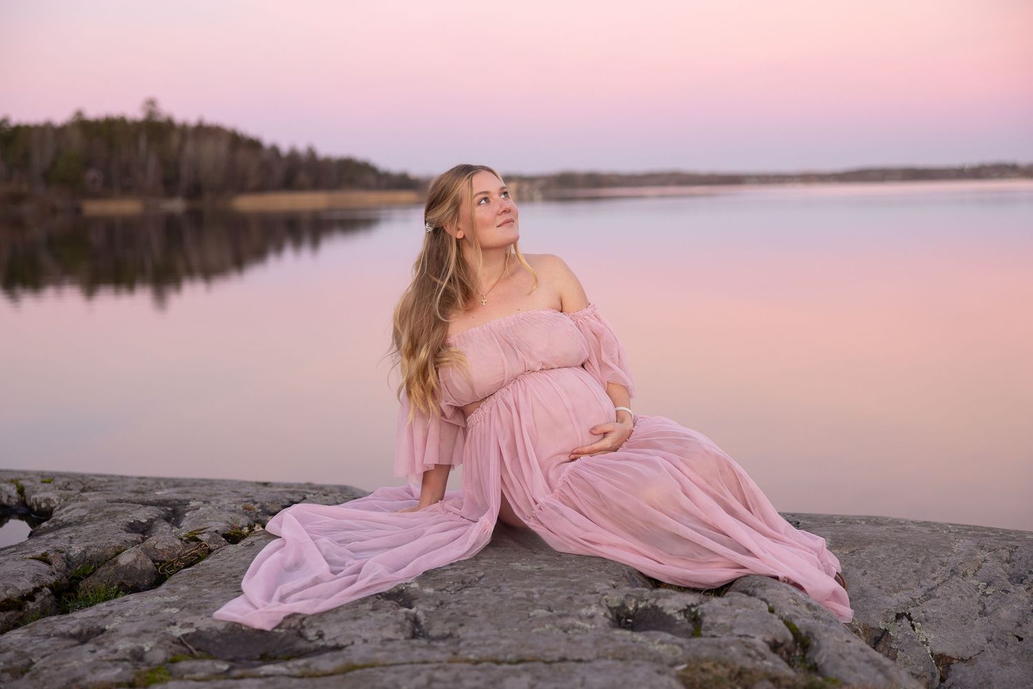 Gravid blondkvinna i solnedgång på klippa med rosa himmel i bakgrunden. Gravid fotografering av emeraldweddings gravidfotograferingdrevviken