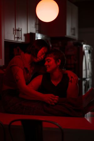 Bathed in red light, a woman sits on her kitchen counter and hugs her partner, while they rest their temples together.