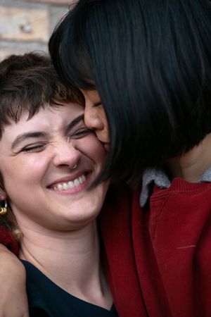Closeup of a woman kissing her partner on the cheek, while her partner scrunches their nose and smiles.