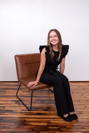 A woman perches on the edge of her chair and smiles at the camera wearing all black for professional, studio headshots.