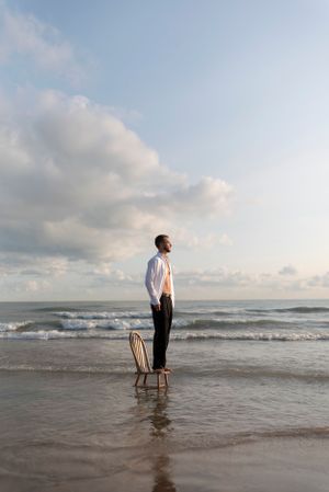 A man in slacks and an unbuttoned shirt stands on a wooden chair placed in the shallow waves on the beach. A warm glow lights him.