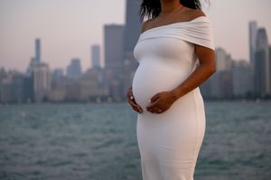 A woman (framed from neck down) wearing a white bardot style bodycon dress holds her pregnant stomach with Chicago and the lake in the bg.