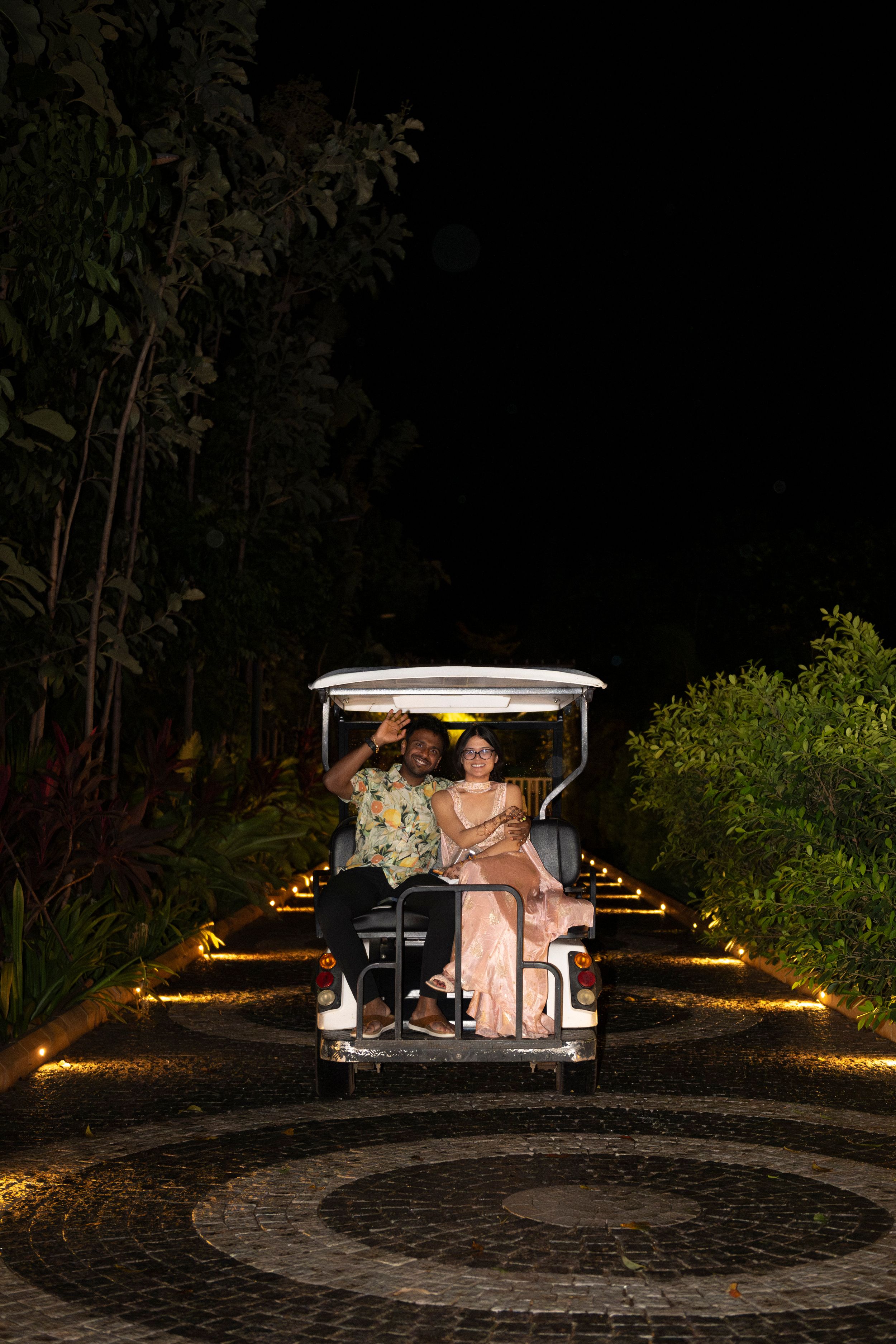 A couple are driven away on the back of a golf cart, waving goodbye at the camera