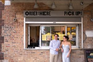 A couple stand, relaxed, in front of an order pick-up window at a taco joint during their welcome party.