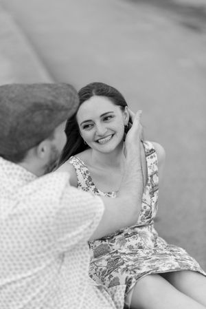 A man seated and facing away from camera, brushes the hair behind his fianceé's ear, while she sits and smiles at him (B&W).